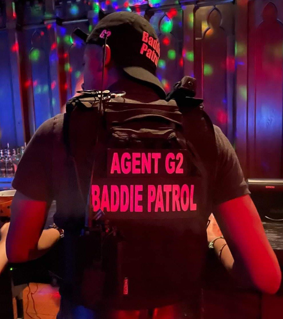 Agent G2 seen from behind in a black Baddie Patrol cap and tactical vest with glowing pink Agent G2 and Baddie Patrol patches, under colorful club lights